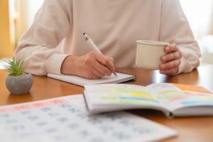 Woman writing notes and managing activities in a planner and calendar, balancing personal and professional life while enjoying coffee at a wooden table in a bright room